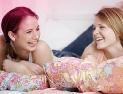 Billede af to unge kvinder liggende i sengen og griner. Billedet viser, at puberteten sker mange ændringer i kroppen, men det er helt normalt.